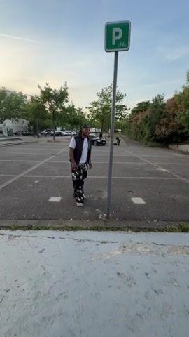 In Saint-Priest, France, an outdoor parking lot is depicted during the late afternoon or early evening under a bright sky. A man with dreadlocks, wearing a white t-shirt, a dark vest, and black and white patterned trousers, walks towards a tall silver pole. The pole features a green sign displaying a white "P" and a bicycle icon, indicating bicycle parking. The man enthusiastically grabs the pole, lifts himself up using only his arms, and then looks upward with an expression of surprise and triumph. In the background, other cars are visible, along with trees and some buildings, suggesting an urban or suburban setting. The man's right arm shows a visible tattoo.