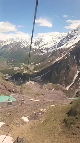 This video shows a view from a cable car descending a mountain. The cable car is moving down a steep slope, with snow-capped mountains in the background. Below, there is a valley with a small village or resort town. There are other cable car cabins visible on the same cable line, some moving in the opposite direction. The terrain is rocky and covered with sparse vegetation in the foreground, with patches of snow. The sky is blue with some white clouds. The camera angle is looking downwards and forwards, offering a panoramic view of the mountainous landscape.