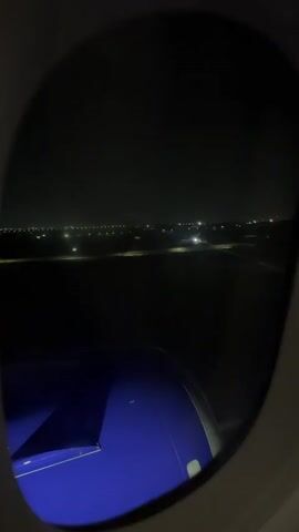 The video shows a view from an airplane window at night. Outside the window, a runway is visible with lights and some distant city lights. The wing of the airplane, which is painted blue and white, is in the foreground. The airplane appears to be either taxiing on the runway or taking off, as the lights on the runway are illuminated and the distant lights are moving.
