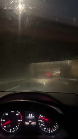 The video shows the interior of a car at night, likely in bad weather. The windshield is wet, and the wipers are active. The dashboard is visible, showing a speedometer with the needle indicating a speed of 53-54 mph. The car is on a road, possibly a highway or tunnel, with other cars visible ahead. The environment appears dark and foggy, with streetlights or headlights illuminating the road.