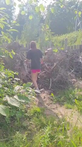 A person is walking up a pile of roots and debris. The person is wearing a dark t-shirt and pink shorts. The person is moving uphill. The background is a fence and trees. The lighting suggests it is daytime.