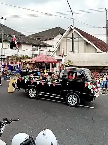 A black pickup truck is driving down a street. The truck is decorated with flags and banners. Several people are riding in the truck bed. In the background, there is a building that appears to be a church, and some tents are set up on the side of the road. There are also some people walking on the sidewalk. The sky is overcast.