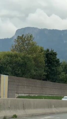 The video is a roadside view from a moving vehicle. A concrete barrier runs along the right side of the frame. Beyond the barrier, there is a sound barrier, followed by trees and then a mountain range covered in greenery and clouds. Several vehicles, including cars and trucks, are visible on the road. The sky is overcast.