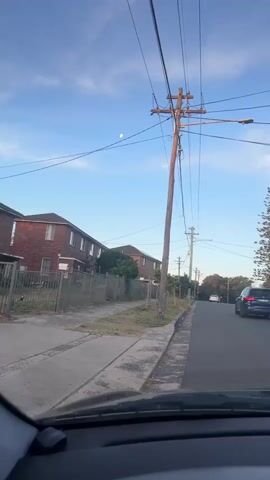 The video, taken from inside a car, shows a residential street on a sunny day.  The car is moving slowly forward on a two-lane asphalt road with a sidewalk on the left. Houses are visible on both sides of the street, mostly brick.  Power lines and wooden utility poles are strung above the street.  Several parked cars line the right side of the road.  A tall, pine-like tree is visible in the distance on the right side of the street. The sky is blue with some scattered clouds, and the moon is visible. The dashboard of the car is visible.