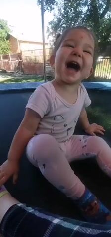 This is a short video clip of a young child with dark hair tied up in two small buns, wearing a light pink onesie with a bear graphic and light pink leggings with a white pattern. The child is sitting on a trampoline in a backyard. The child is laughing heartily and looking towards the camera. The setting appears to be outdoors during the daytime, with sunlight filtering through trees in the background. A wooden fence and part of a building are visible behind the trampoline. The child's expression is one of pure joy and amusement. The overall mood of the video is happy and playful. There is no visible text in the clip.