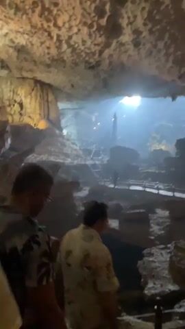 The video shows a group of people walking through a large, illuminated cave. The cave walls and ceiling are covered in stalactites and stalagmites, creating a textured, natural environment. Wooden walkways with railings guide the visitors through the cave, indicating it is a tourist attraction. Sunlight streams in from an opening in the cave ceiling, creating a dramatic spotlight effect on a central rock formation. Several people are visible walking on the path, some in groups, others individually. The visitors appear to be dressed in casual attire, suggesting a warm climate. The overall atmosphere is one of exploration and wonder, with the natural beauty of the cave enhanced by artificial lighting.