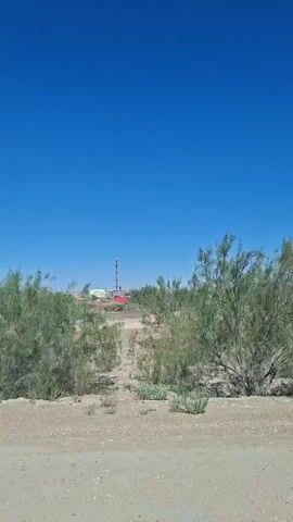 The video shows a view from a vehicle traveling on a dirt road. The sky is clear and blue. On either side of the road are sparse, green, bushy trees and dry, sandy ground. In the distance, there appears to be some industrial structures, including a tall tower and some red buildings. The camera pans slightly to show more of the landscape and the road ahead. The vehicle's side mirror is visible in one shot, reflecting the passing scenery.
