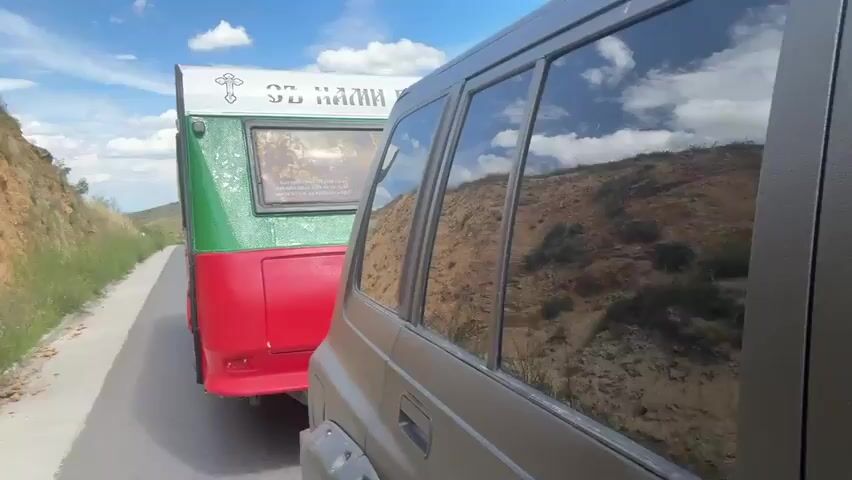 The video shows a side view of a dark-colored SUV from the perspective of someone sitting in the driver's seat. The SUV is parked on the side of a paved road, which runs alongside a steep, rocky, and vegetated hillside on the left. In front of the SUV, also on the road, is a trailer. The trailer is painted green and red and has a white top section with a cross and some text. A rectangular panel on the trailer appears to display an image or artwork. The windows of the SUV reflect the sky, clouds, and the surrounding rocky landscape. The sun is shining, and the sky is blue with scattered white clouds. The overall scene suggests a rural or roadside setting.