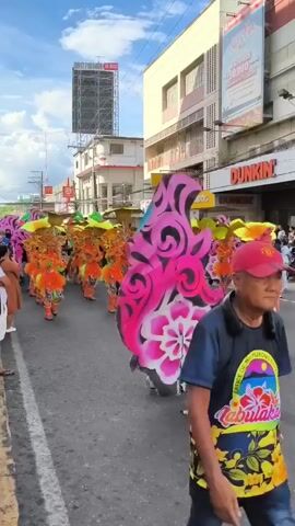 The video shows a street parade. A large group of people, all wearing orange fringed costumes and large, yellow flower-shaped hats, are marching down the road. A person carries a large, flat flower prop with pink and black details in front of the group. Businesses like Dunkin' are visible along the street. A tall billboard with a phone number is on the left side of the street. The sky is blue with some clouds. People stand along the side of the road, watching the parade.
