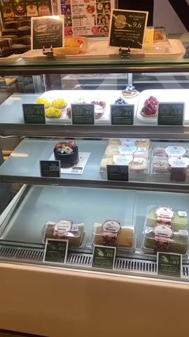 The video shows a close-up view of a refrigerated display case filled with various pastries and cakes in a bakery. The display case is divided into multiple shelves. On the top shelf, there are several small tarts and cakes with fruit toppings, as well as some larger pastries with what appears to be jam filling. Some of these items have small price tags in front of them. The middle shelf contains individually packaged cakes and pastries, including what look like chocolate cakes, donuts, and possibly some kind of layered cake or pastry. Some of these items also have price tags and labels. The bottom shelf features several Swiss rolls, some with green filling and others with a more traditional yellow or cream filling, all individually packaged and labeled with prices. The lighting in the bakery is bright, illuminating the display of desserts. The overall environment appears clean and well-maintained. There are no people visible in the video.