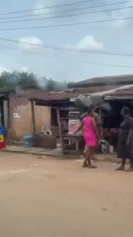 The video, shot from inside a moving vehicle, depicts a street scene in what appears to be a developing country. The road is unpaved and dusty. Several small, makeshift shops or stalls line the street, some with corrugated metal roofs. One stall has a sign that reads "GIFT SHOP". A yellow auto-rickshaw is parked near one of the structures. There are stacked crates in blue, yellow, and red. Power lines are visible overhead. As the vehicle moves, a man in a white garment walks by. A motorcycle carrying two people, a man in a navy shirt and a woman in a blue and orange patterned dress, passes the vehicle. The video continues to show the structures lining the street, some with tiled walls.  A water tower with black and blue tanks is visible in the background.  The overall impression is of a modest, perhaps rural or semi-urban environment.