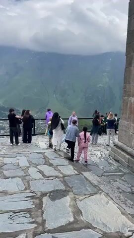 A group of people are gathered on a stone patio overlooking a vast, green mountain range. A portion of a stone building is visible on the right side of the frame. Several individuals are taking pictures of a group of children dressed in traditional green clothing, some of whom are holding instruments. The children appear to be posing for a photograph. The sky is cloudy.