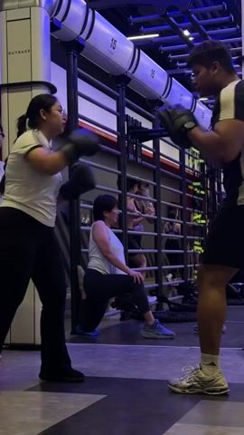 The video shows a gym where two people are practicing boxing. In the foreground, a woman wearing a white shirt and black leggings is sparring with a man wearing a black t-shirt and black shorts. They are both wearing boxing gloves. The woman is throwing a punch, and the man is holding pads to block it. In the background, a woman in a light blue top and black pants is performing a lunge exercise. Other people can be seen in the background engaged in various exercises. The gym appears to be modern with metal racks and blue and white lighting.