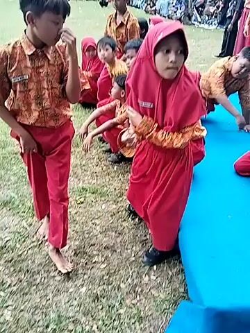 The video shows a group of young children standing and sitting on a grassy field. Some are wearing matching outfits of red pants and batik-style shirts, while others are wearing red dresses and headscarves. One child is barefoot. Some children are standing and walking around, while others are sitting. Behind them, there is a blue inflatable object. In the background, there are trees, a red and white flag, and more people. The camera moves slightly, focusing on different children in the group.