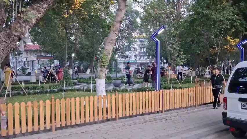 The video shows a park scene with people walking and standing. In the foreground, a woman in a red and white floral dress walks to the right, holding a yellow and orange toy. A young child, possibly a girl, walks beside her, holding her hand. To the right of them, another woman in black athletic wear walks towards the left. A low, light-colored wooden picket fence runs along the edge of the park. Behind the fence, there is a well-maintained green lawn with some small trees and shrubs. Modern blue-and-white streetlights are visible in the park. In the background, more people are scattered throughout the park, and buildings can be seen beyond the trees. A white van or SUV is parked on the right side of the frame. The lighting suggests it is daytime, possibly late afternoon or early evening.