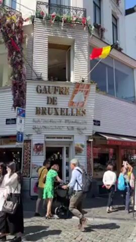 The video shows the exterior of "Gaufre de Bruxelles," a waffle shop. People are walking on the cobblestone street in front of the shop. Some people are entering the shop. The shop is painted white and has gold lettering. A Belgian flag is flying. The shop is decorated with flowers.  There are other shops adjacent to the waffle shop.