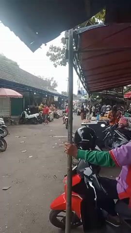 A busy outdoor market scene with a focus on a street in front of stalls. The street is paved with dirt or gravel and is lined with buildings or stalls on both sides. A red motorcycle is in the foreground, with a person in a purple and green shirt holding onto its handlebars. Several other motorcycles are parked or moving around the market. People are walking and riding motorcycles, and many stalls are visible, displaying goods. The overall atmosphere appears to be one of active commerce and a bustling public space.