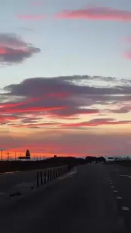 The video shows a view from a car on a highway at sunset. The sky is a mix of orange, pink, and grey clouds. On the side of the highway, there are utility poles, a black metal fence, and white and black traffic barriers. A large road sign is also visible. There are a few other cars on the highway in the distance.