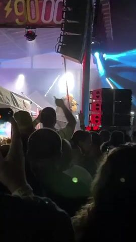 The video shows a live performance at an outdoor venue. The stage is lit with bright lights, and there are speakers visible on the right side of the frame. In the foreground, the silhouettes of a crowd are visible, with some hands raised. On stage, two men are performing. One man, who appears to be the main performer, is bald, wearing a dark jacket, and singing into a microphone. He is facing away from the camera. To his right, another man, wearing a baseball cap and a t-shirt with a graphic design, is also singing and gesturing. He is facing the crowd. There are some pyrotechnic effects on stage, appearing as streaks of light. The overall atmosphere appears to be energetic and engaging for the audience.