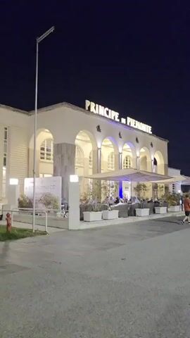 The video displays a long shot of the exterior of a building at night. The building is white with a series of arches running across its front. Above the arches, in large illuminated letters, is the text "PRINCIPE DI PIEMONTE". Below the arches, there is an outdoor seating area with tables and chairs, and several people are seated at the tables, appearing to be dining or socializing. Large white umbrellas are set up over some of the seating areas. In front of the building, there are planters with greenery and a sidewalk. A tall lamppost stands to the left of the building. The overall atmosphere is that of an upscale outdoor restaurant or cafe.