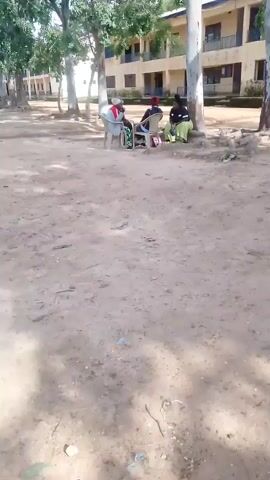 The video shows a dirt ground in the foreground with some debris scattered. In the background, three people are sitting on chairs under trees. Two of the people are women, and one appears to be a man. They are seated in a semi-circle and appear to be engaged in conversation. Behind them is a multi-story building with windows. The overall environment appears to be outdoors, possibly on a school campus or a similar public space.