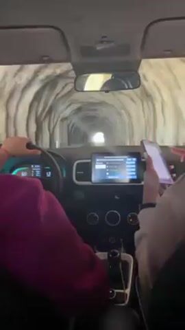 From inside a vehicle, a view shows the car driving through a tunnel. The dashboard and steering wheel are visible. The driver, on the left, wears a pink sweatshirt and has both hands on the wheel. The passenger, on the right, has one hand on a phone held upright. The vehicle's digital display screen is lit. The interior of the tunnel has a rough, rocky appearance with a light source at the end.