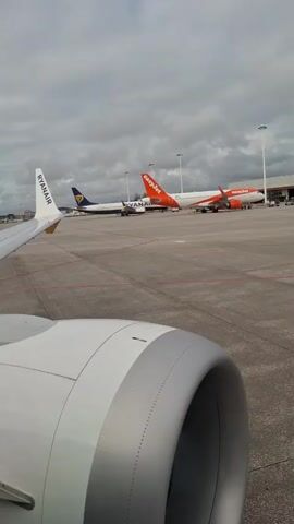 The video shows a view from inside an airplane, looking out over the wing and engine toward the airport tarmac. Two Ryanair planes and two EasyJet planes are parked on the tarmac under a cloudy sky. The engine is white with a gray accent, and the Ryanair wing is visible on the left.