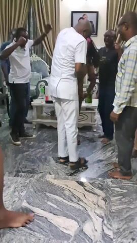 The video shows a group of people dancing in what appears to be a living room.  There are about 7-8 people visible, including one person in a white suit and sandals who seems to be leading the dancing. Others are dressed in casual clothes like jeans, plaid shirts, and a yellow top. Some people are barefoot, and one person is only visible from the knees down, with purple nail polish. The room is decorated with marble flooring, cream-colored curtains, a white couch, and a small table with bottles on it. There's also a framed picture on the wall behind the group. The atmosphere seems celebratory and festive.