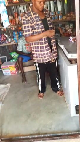 A person in a plaid shirt and track pants stands in the doorway of a small store, holding a phone. The store has shelves stocked with items. A second person, wearing a striped shirt and blue shorts, stands behind them. There are bags of snacks hanging on the side of the doorway. A box of Indomie noodles is visible on the floor, along with several onions and two pairs of sandals.