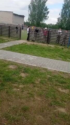 The video shows an outdoor scene on a grassy field with a grey brick path leading to a wooden fence. Beyond the fence, a group of people is visible, some are holding what appear to be sticks or poles. To the left is a simple building with a flat roof. The sky is overcast. The camera is shaky and handheld, giving a perspective from ground level.