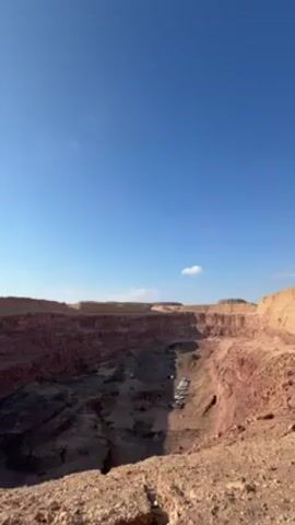The video shows a wide shot of a large, deep canyon under a clear blue sky with sparse clouds. The canyon walls are layered with reddish-brown rock formations. At the bottom of the canyon, several white vehicles are visible, suggesting human activity. The surrounding landscape appears arid and desert-like, with rocky terrain. The camera remains still throughout the video.