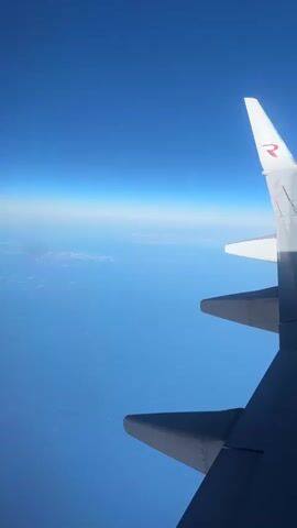 The video captures a view from an airplane window looking out at a clear blue sky. The wing of the airplane is prominently in the foreground, extending from the right side of the frame towards the bottom. The wing is dark grey on the lower portion and transitions to white towards the tip. A red logo, possibly an 'R', is visible on the white tip of the wing. Below the wing, faint clouds or landmass can be seen in the distance, blending with the horizon. The overall environment is vast and open, suggesting the airplane is flying at a high altitude over a clear day.