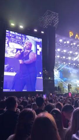 This video shows a concert at night, indicated by the dark sky and the stage lights. A large crowd of people is gathered in front of a brightly lit stage. Above the stage, the words "PARK LIVE" are displayed in large, illuminated letters. The stage features large LED screens displaying images of performers, and the lighting is dynamic with beams of light and fog effects. Many people in the crowd are holding up their phones, presumably to record the performance. The overall atmosphere is one of excitement and engagement with the event.