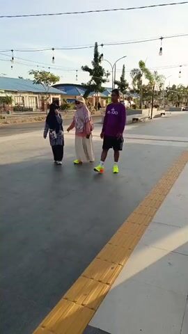 Three people are walking on a paved walkway. The person on the left is wearing a blue headscarf and a dark blue and white patterned dress. The person in the middle is wearing a light pink headscarf and a cream-colored skirt. The person on the right is wearing a purple long-sleeved shirt and black shorts. They are walking away from the camera, towards a building in the background. The sky is clear and the sun is shining. There are lights strung across the walkway.