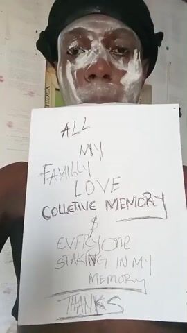 A person with dark skin and white paste on their face and wearing a black cap is holding a white sheet of paper with handwritten text. The text reads "ALL MY FAMILY LOVE COLLECTIVE MEMORY & EVERYONE STAKING IN MY MEMORY THANKS."  A book titled "CHIDEX" is visible in the background. The background appears to be an indoor wall with light patterns.