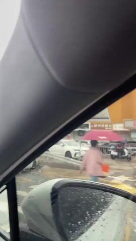 The video shows a street scene from inside a vehicle. It is raining, and the windshield has raindrops on it. Several cars are visible, including white sedans and SUVs, and some red cars parked further down the street. A white ramp or metal sheet is positioned across the road, with a white car driving over it. Buildings line the street, some with signs in Chinese. The sky is overcast. The video appears to be a continuous shot without any editing.