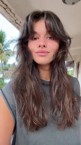 A young woman stands on a porch or balcony, facing the camera. She is wearing a gray t-shirt and has long, layered brown hair with bangs. She has a nose ring and is smiling. In the background, there are palm trees and other greenery. She pouts her lips towards the camera and then smiles.