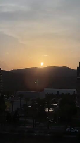 A time-lapse video captures the sun setting behind a silhouetted mountain range. The sun, a bright orb, is partially obscured by the mountain, casting warm orange and yellow hues across the sky. A crescent-shaped shadow, indicating a solar eclipse, is visible against the sun's disk. Below the mountain range lies a darkened landscape with the outlines of buildings, palm trees, and streetlights. The sky transitions from a bright orange near the horizon to a lighter grey towards the top. The video appears to be filmed from a high vantage point, possibly a building, overlooking the city and surrounding natural landscape.