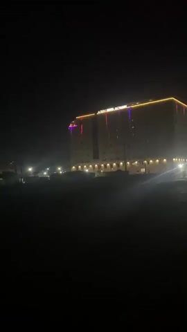 A low-angle night shot captures a large, multi-story hotel building. The building's facade is illuminated by a strip of yellow lights running along the top edge, with vertical accents of pink and purple lights on the left side. Additional warm lighting emanates from the ground floor, suggesting an entrance or lobby area. A few parked cars are visible in front of the building, their headlights casting a soft glow. The foreground is dark and indistinct, possibly representing unpaved ground or a field. The overall atmosphere is somewhat hazy, potentially due to atmospheric conditions or artificial lighting effects. The video is extremely short, lasting only four seconds, with no discernible action or interaction.