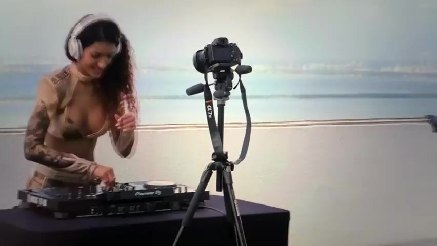 This media file captures a woman DJing in a contemporary setting. She is the central subject, wearing headphones and a stylish, revealing outfit. In front of her is a DJ setup, including a Pioneer DJ turntable and mixer. A camera on a tripod is positioned to record her performance, suggesting a live stream or a recorded session.

The scene appears to be outdoors, likely on a balcony or rooftop, with a blurred cityscape and a body of water visible in the background. The sky is overcast, suggesting a diffused natural light, and the overall atmosphere is modern and sleek. The woman's expression is one of concentration and enjoyment as she interacts with the DJ equipment.

There is no explicit indication of the time of day or specific weather conditions beyond the overcast sky. The location context provided places this in Falenty Nowe, Poland. The visible text on the DJ equipment is "Pioneer DJ".