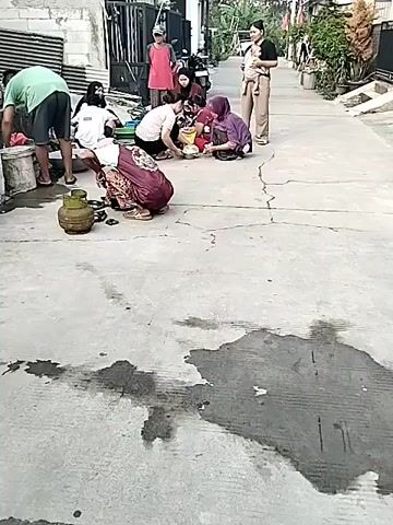 A group of people are gathered on a paved outdoor area that appears to be a narrow alley or street. Several women and a man are crouched or kneeling on the ground, engaged in an activity. One woman in the foreground, wearing a patterned maroon and white skirt and a dark headscarf, is interacting with a green gas cylinder and some small items on the ground. To her left, a man in a green t-shirt and dark shorts is also crouched near some containers. Other women are gathered around a central area, some of them appearing to be washing or preparing something. One woman is standing and holding a baby. The environment includes buildings with corrugated metal siding and a motorcycle parked in the background. The ground is cracked concrete with several wet patches.