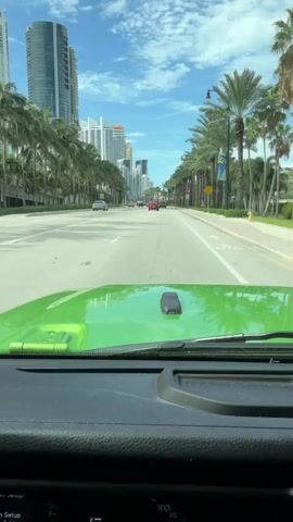 The video shows a first-person perspective of someone driving a bright green Jeep down a wide, paved road lined with tall palm trees on both sides. In the distance, a cityscape with numerous high-rise buildings is visible under a blue sky with scattered clouds. Cars are driving in the same direction, and some are seen on the opposite side of the road. The road is marked with white lines, and the overall scene suggests a sunny day in a coastal urban environment.