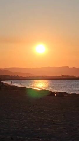 A video of a beach at sunset shows the sun setting over the ocean, casting a warm glow on the water and the sand. The water is calm, with gentle waves lapping at the shore. The sky is a clear orange, with a few clouds scattered across the horizon. In the distance, a range of mountains can be seen silhouetted against the setting sun. There are a few people on the beach, walking along the shore and enjoying the view.