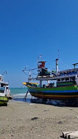 The video shows a coastal area with several boats on the shore. The first boat, prominent in the initial frames, is a colorful fishing vessel, green, blue, and yellow, supported by sandbags and resting on the beach. It appears to be undergoing repairs or maintenance, with visible scaffolding and ropes. The background reveals a boatyard or repair facility with more wooden structures, tarps, and building materials. Other boats are visible in various states of construction or repair, some partially covered. The ground is a mix of sand and debris, including scattered wood, plastic, and general refuse. The sky is clear and blue, indicating a sunny day. The overall scene depicts a working environment for boat construction and repair on a beach.