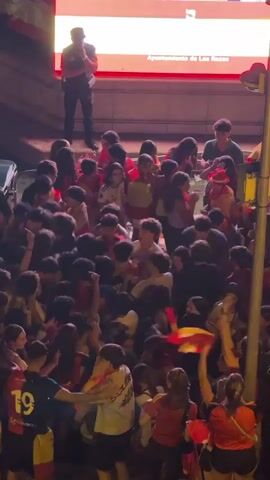 The video shows a large crowd of young people, mostly teenagers and young adults, gathered outdoors at night. They are wearing red and yellow clothing, and many are holding and waving Spanish flags. The crowd appears to be excited and celebratory. There is a large LED screen in the background with text that is partially visible. There are also streetlights and some police officers present. The overall impression is a public gathering celebrating a national event or victory.