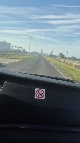 The view from inside the dashboard of a car is seen driving on a straight road.  The sky above is light blue with thin, high clouds. On the left side of the road is a large white tank with various metal pipes and structures around it. Street lamps line both sides of the road, and beyond a small grass strip on the right side are train tracks.  Further down the road, more industrial-looking buildings can be seen, along with a passing truck. A "no smoking" sign is affixed to the dashboard in the foreground, and a Dominican Republic flag hangs from the rearview mirror.