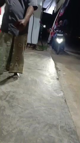 The video shows a street scene at night. In the foreground, a person's lower body is visible from the waist down, wearing a patterned skirt and sandals. They are standing on a concrete surface. In the background, a motorcycle with its headlights on is approaching. Two people are riding the motorcycle, one driving and one passenger. They are illuminated by the headlights and appear to be moving towards the viewer. Flags are visible attached to a pole behind the motorcycle. The environment appears to be outdoors, possibly a roadside or a small business area, with some structures and hanging items in the background. The lighting is dim, with the motorcycle's headlights being the primary light source in the background.