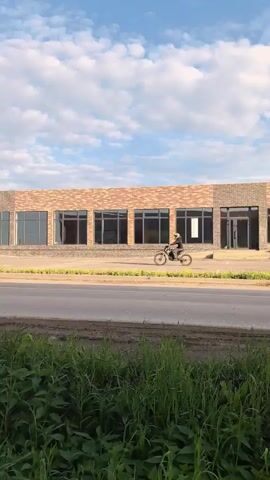 This is a vertical video clip showing a person riding a dirt bike in front of a brick building with large windows. The rider is wearing a helmet and appears to be performing a wheelie, with the front wheel of the bike lifted off the ground. The building is modern in design and has a light brown brick facade. The windows are large and dark, reflecting the sky.

In the foreground, there is a patch of green grass. The road in front of the building is paved. The sky is partly cloudy with a mix of blue sky and white clouds. The lighting suggests it is daytime, likely late afternoon or early evening, given the soft light. The scene appears to be outdoors, possibly in an industrial or commercial area, given the building style. The overall mood is dynamic and energetic due to the action of the dirt bike rider. There is no visible text in the clip. The location is specified as Gorniy, Russia.
