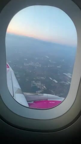 The video shows a view from an airplane window looking out at the landscape below during what appears to be sunrise or sunset. The wing of the airplane, which is white with pink accents, is visible on the left side of the frame. The engine, also featuring pink branding, is partially visible. Below the aircraft, a sprawling urban or semi-urban area can be seen with buildings, roads, and patches of green land. The sky is a gradient of blue, with soft hues of pink and orange visible near the horizon, indicating twilight. The overall impression is one of travel and the scenic beauty of looking down on the Earth from high above.