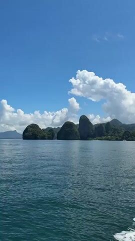 The video captures a scenic view of limestone karst islands emerging from the tranquil blue ocean under a clear sky filled with scattered white clouds. The perspective suggests the viewer is on a moving boat, as evidenced by the white wake trailing at the bottom right of the frame. The islands are lush with green vegetation, and their rugged, rocky formations dominate the horizon. The lighting indicates it is daytime, likely late morning or early afternoon, given the bright sunlight and the clarity of the shadows. The overall impression is one of natural beauty and a peaceful, tropical setting.